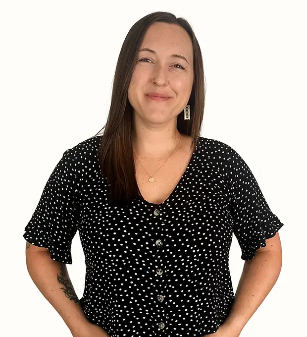 A woman with straight brown hair, wearing a black polka dot blouse, stands smiling with her hands on her hips against a plain white background.